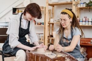 young female master of pottery consulting girl in workwear sitting by wheel.jpg